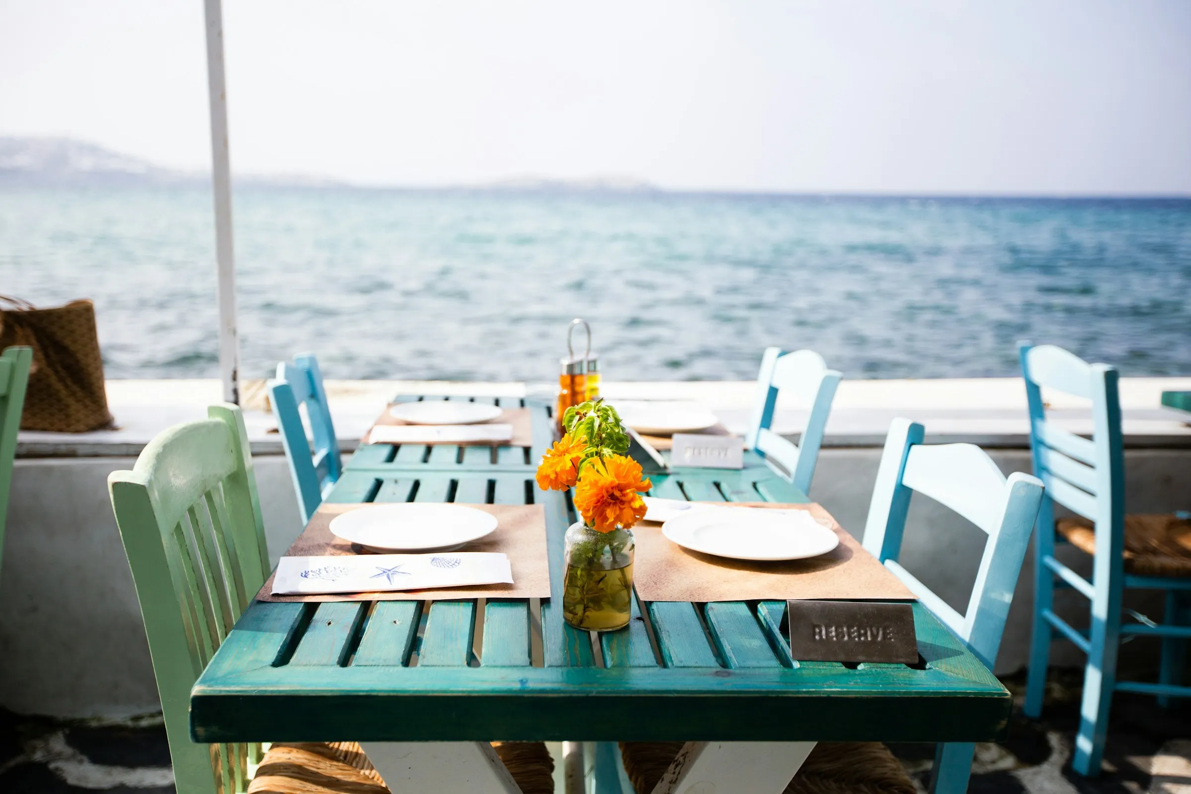 A beachfront restaurant table set for two with a 'RESERVE' sign, blue chairs and ocean view.
